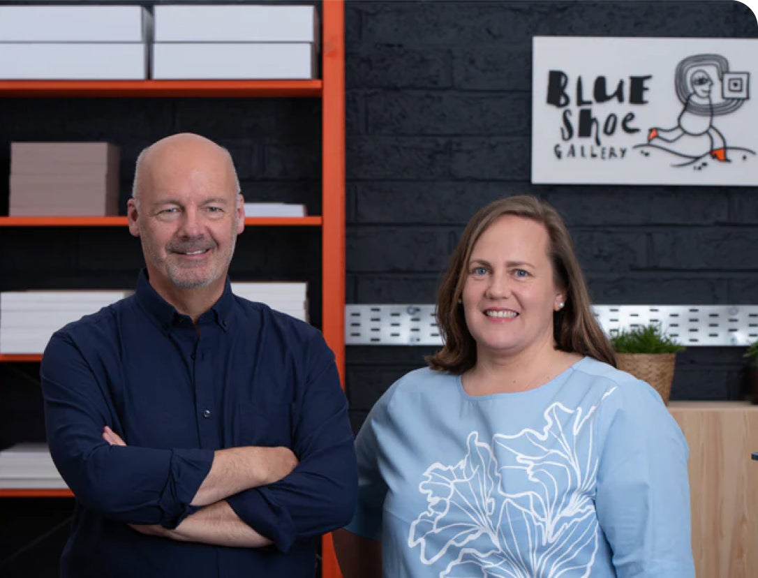 Two people standing in front of a shelf with a sign that reads 'Blue Shoe Gallery'.